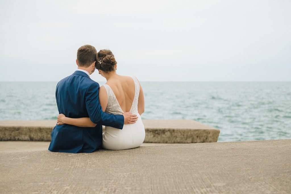 A bride and groom sit together on a ledge overlooking the lake in Chicago during wedding photos with Ed and Aileen Photography. 