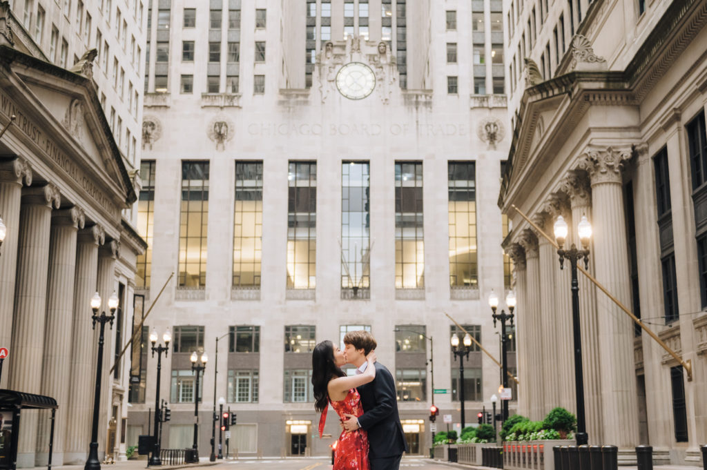 An engaged couple embrace and kiss in front of the impressive buildings in the financial district on the Chicago Loop. 