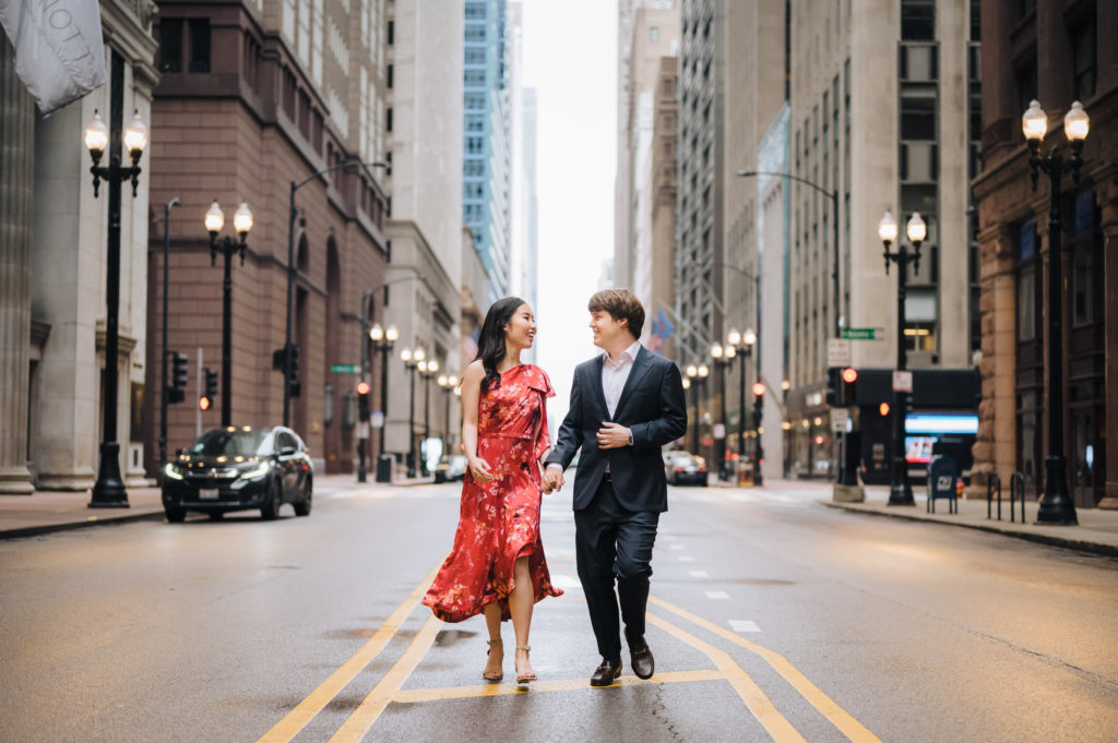 A couple smile at each other while walking hand-in-hand down the middle of a street in Chicago's financial district, one of many unique Chicago photo spots, during an engagement shoot with Ed and Aileen Photography.